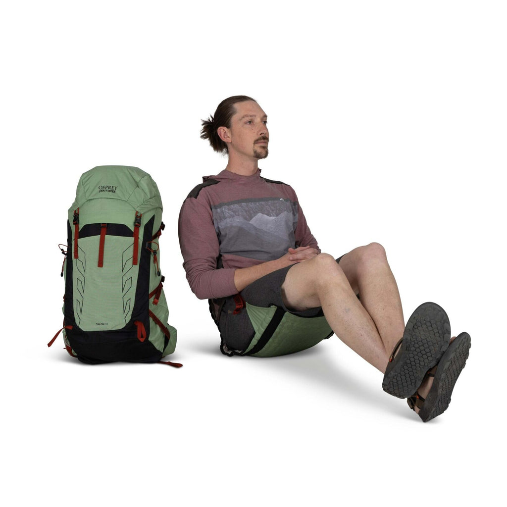 A person sits next to a large light green and black Osprey backpack, wearing a red and gray shirt and shorts, with rugged sandals.