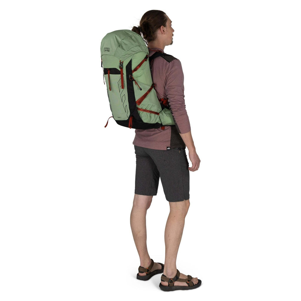 A person stands outdoors wearing a light green backpack, gray shorts, and sandals, facing slightly away from the camera.