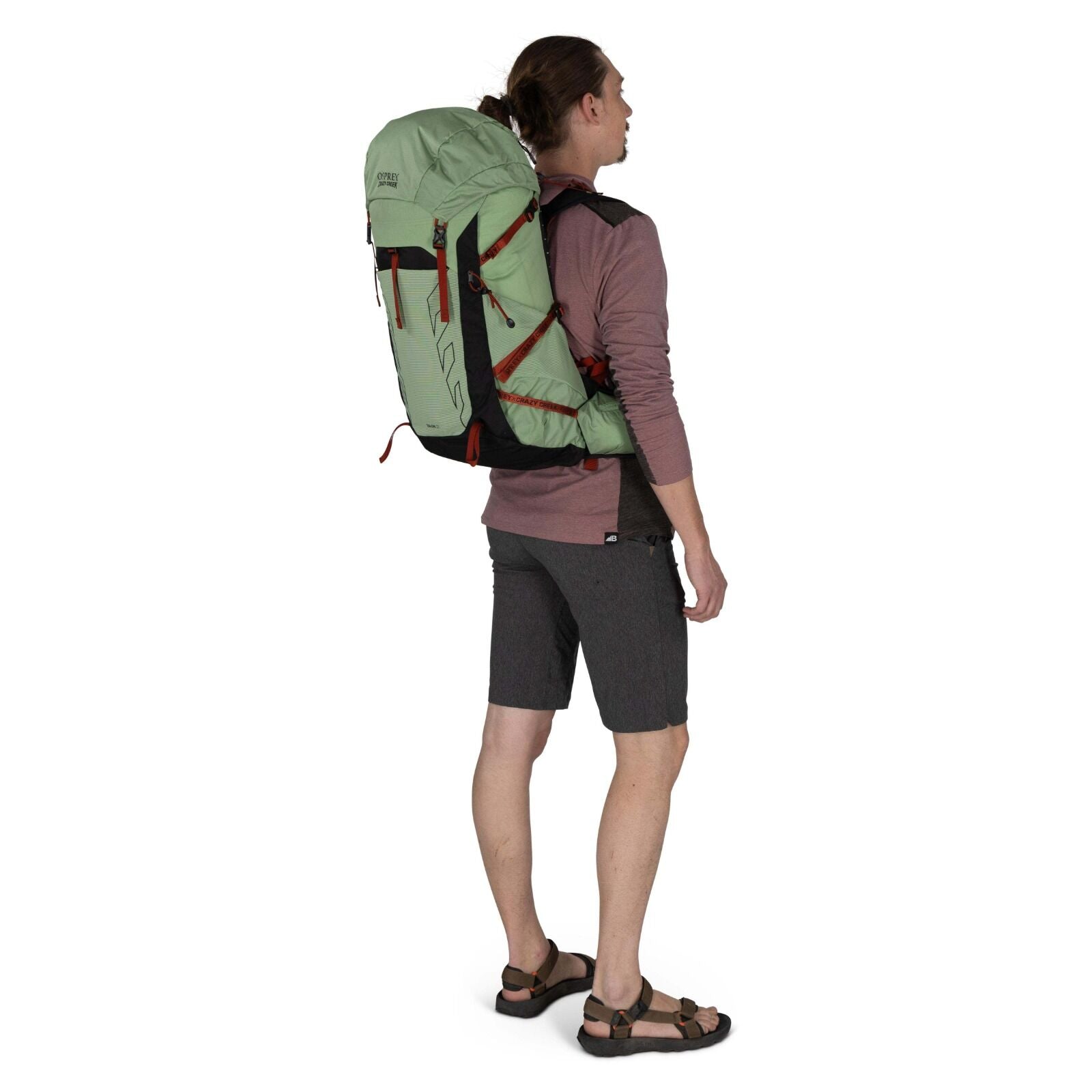 A person stands outdoors wearing a light green backpack, gray shorts, and sandals, facing slightly away from the camera.