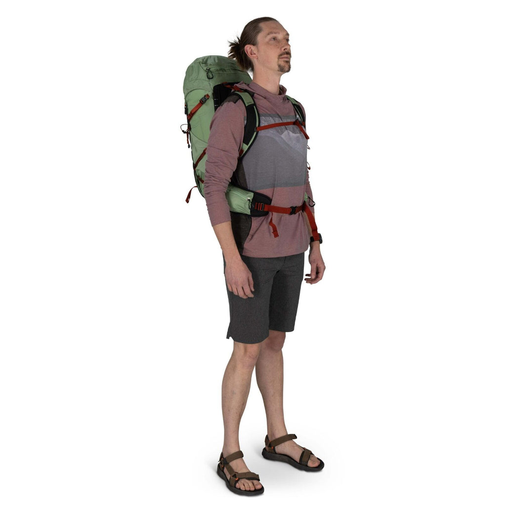 A person stands upright wearing a green backpack, a maroon hoodie, and shorts, ready for an outdoor adventure.